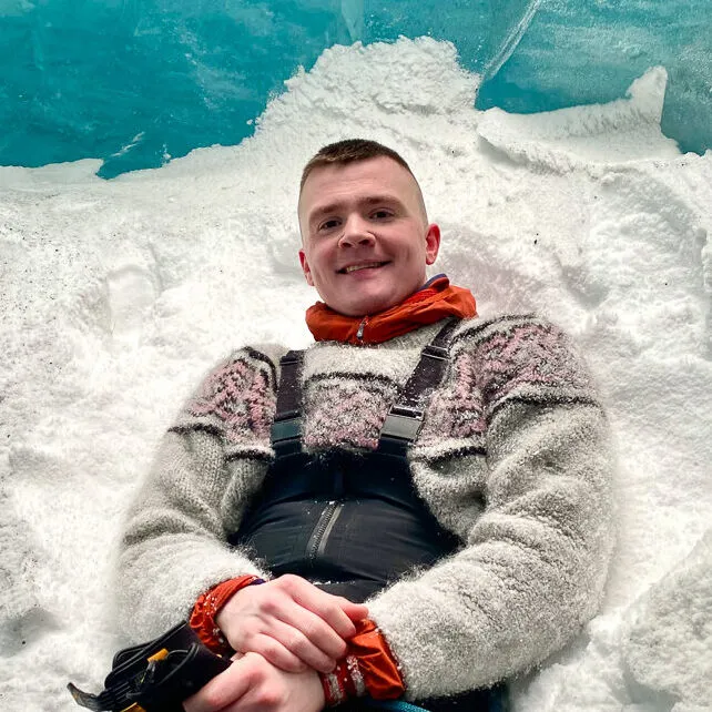 Man wearing an Icelandic wool sweater sits in a pile of snow in front of a blue ice glacier wall.
