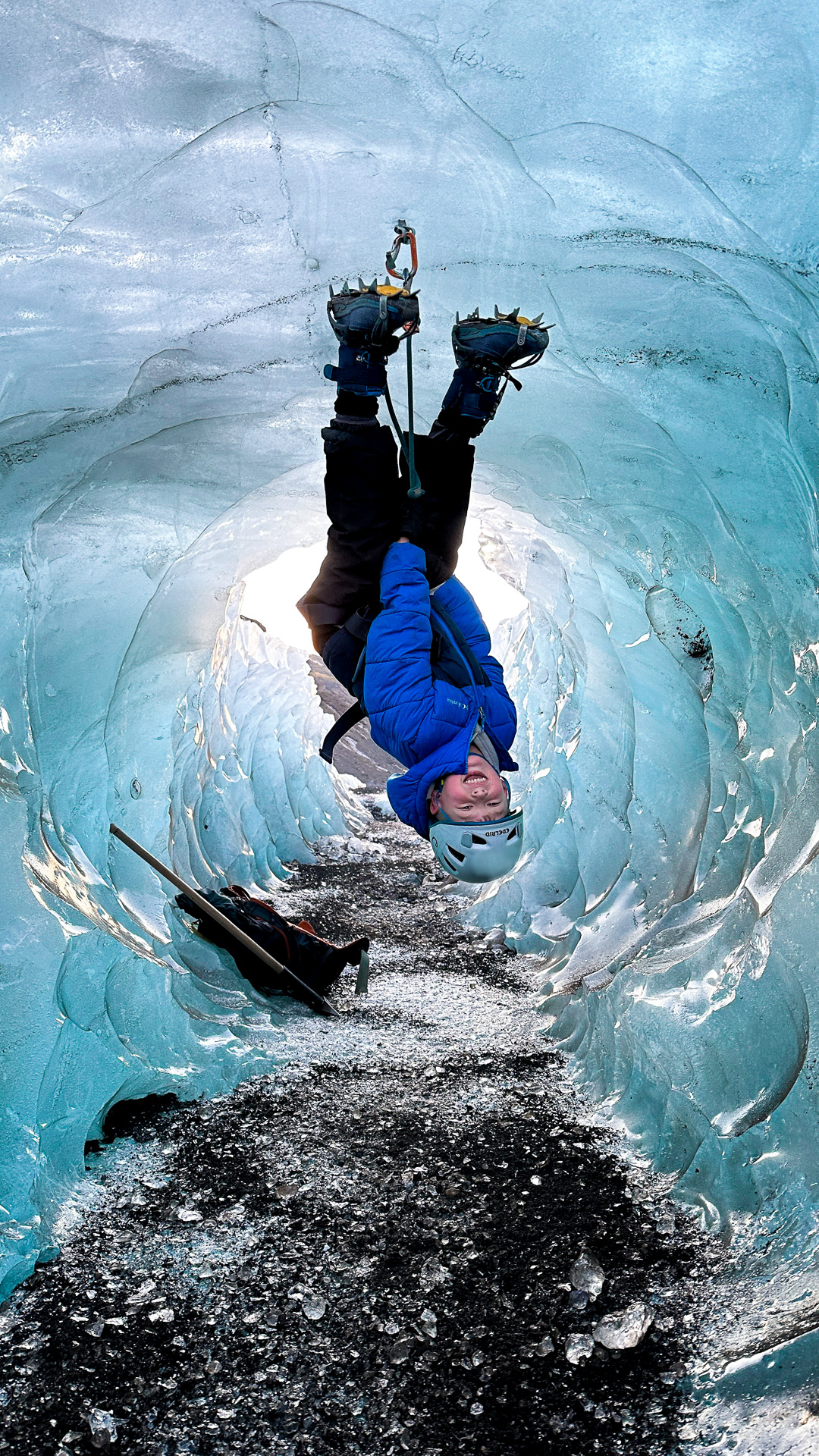 Kid in a blue jacket and helmet hanging upside down from a rope in a blue ice cave.