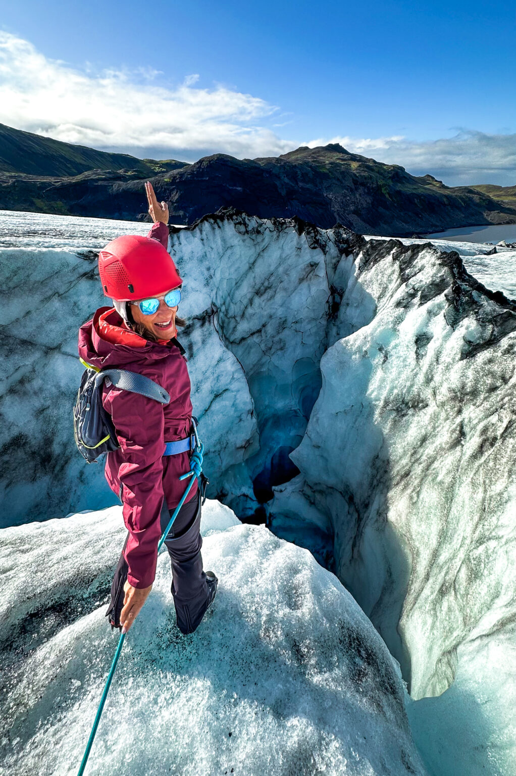 Woman attached to a rope stands at the edge of a deep hole on the glacier with her arm raised on a sunny day with cliffs in the background.